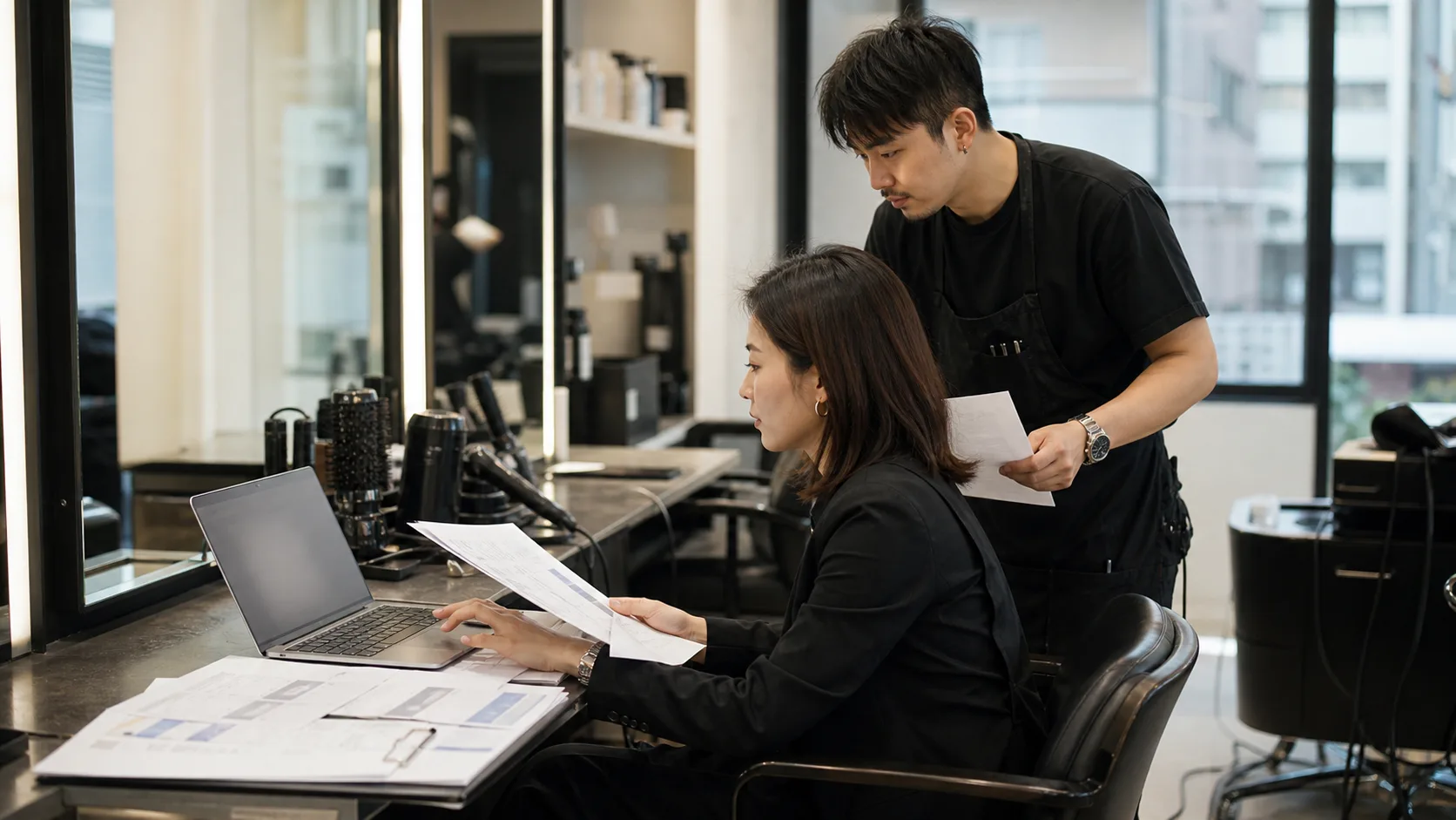 台灣髮廊工作站旁，成年店長與設計師側身核對廣告設定與落地頁資料
